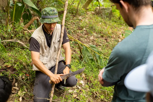 A guide crouching on a forest trail demonstrates a machete to a participant