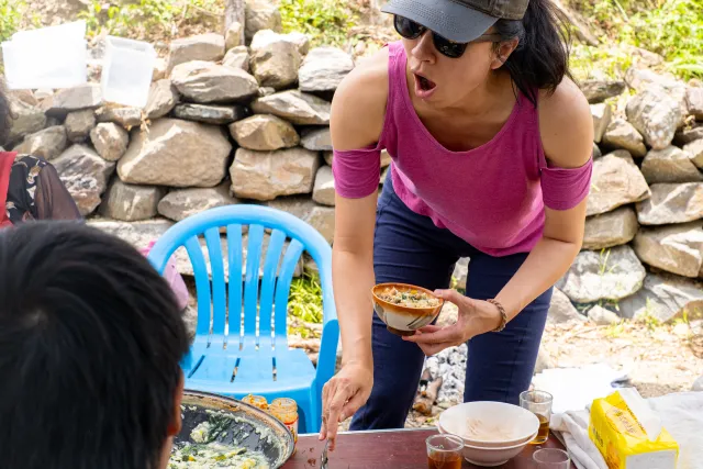 A woman in a pink top and visor leans over an outdoor table, holding a bowl of food with an expression of surprise or delight