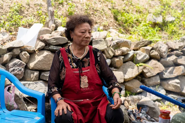 An elderly woman in a red apron sits quietly beside a stone wall outdoors