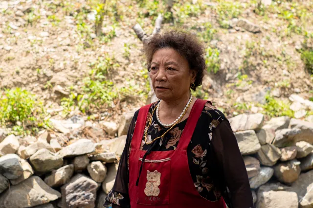 An older indigenous woman in a red apron stands outdoors beside a stone wall, looking solemnly into the distance