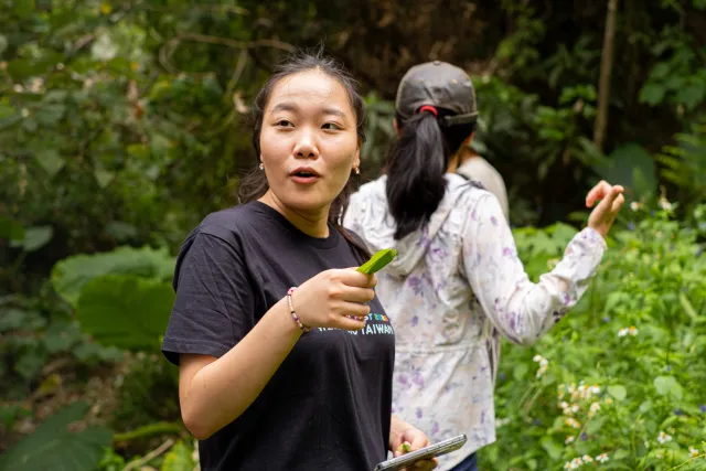 A woman holding a plant stem speaks expressively on a forested trail while another participant examines nearby foliage