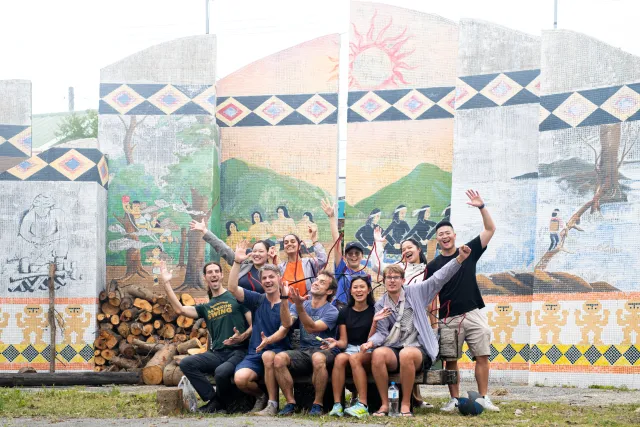 A group of ten smiling visitors pose together in front of a large colorful indigenous mosaic mural