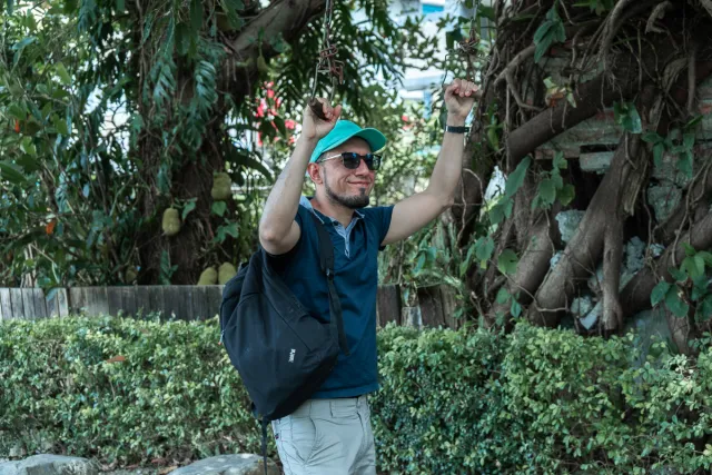 A smiling man in a teal cap and sunglasses grips hanging tree branches outdoors