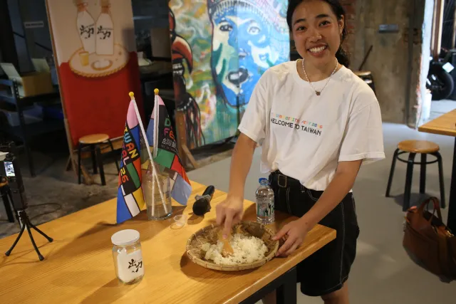 A smiling young woman in a "Welcome to Taiwan" T-shirt kneads cooked rice in a woven tray during a brewing workshop